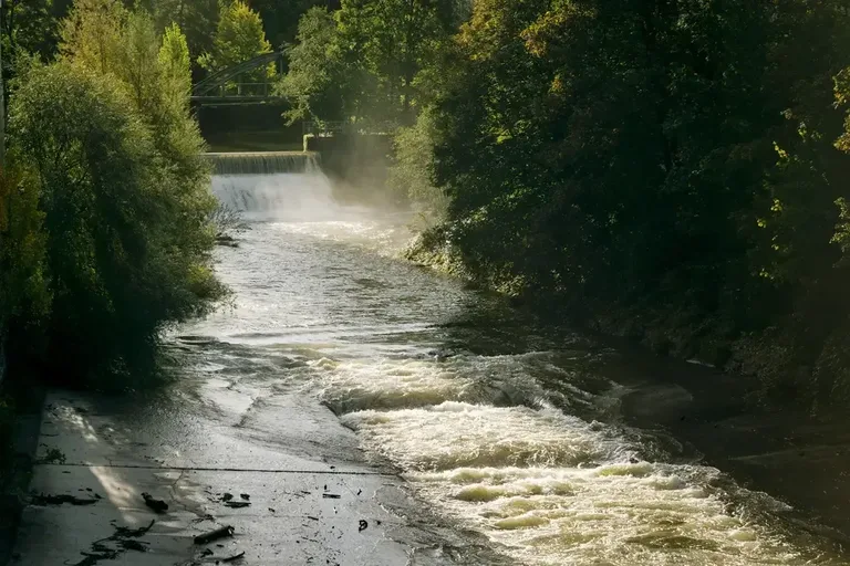 Flusslauf der Töss beim Vitus-Areal Winterthur mit Wasserfall, Ufervegetation und Sonnenlicht im Hintergrund.