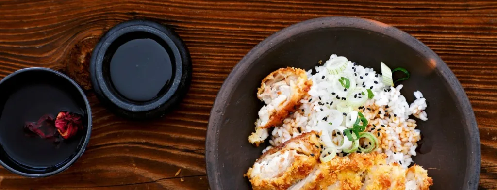 Schüssel mit knusprig paniertem Hühnchen, Reis und Frühlingszwiebeln, daneben zwei kleine Schalen mit Sauce auf einem Holztisch.